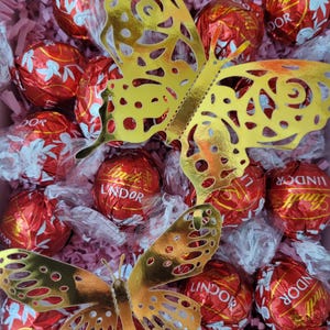 Puede incluir: Una caja de regalo llena de trufas de chocolate Lindor envueltas en papel de aluminio rojo y dos recortes de mariposas doradas. Las mariposas tienen intrincados dise&ntilde;os de alas y est&aacute;n colocadas sobre los chocolates y el relleno de papel rosa.