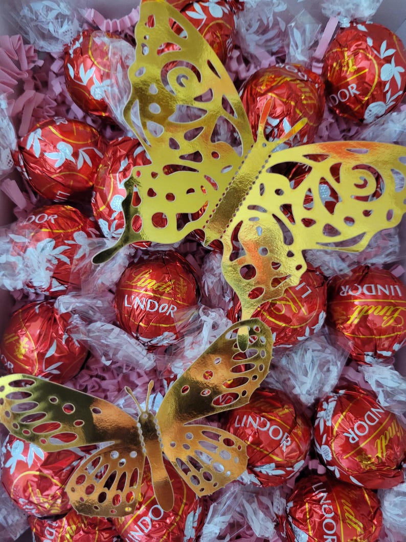 Puede incluir: Una caja de regalo llena de trufas de chocolate Lindor envueltas en papel de aluminio rojo y dos recortes de mariposas doradas. Las mariposas tienen intrincados dise&ntilde;os de alas y est&aacute;n colocadas sobre los chocolates y el relleno de papel rosa.
