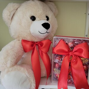 Puede incluir: Un oso de peluche color crema con un lazo de sat&eacute;n rojo alrededor del cuello. Junto al oso hay una caja transparente llena de bombones y un gran lazo de sat&eacute;n rojo. La caja contiene decoraciones de mariposas doradas.