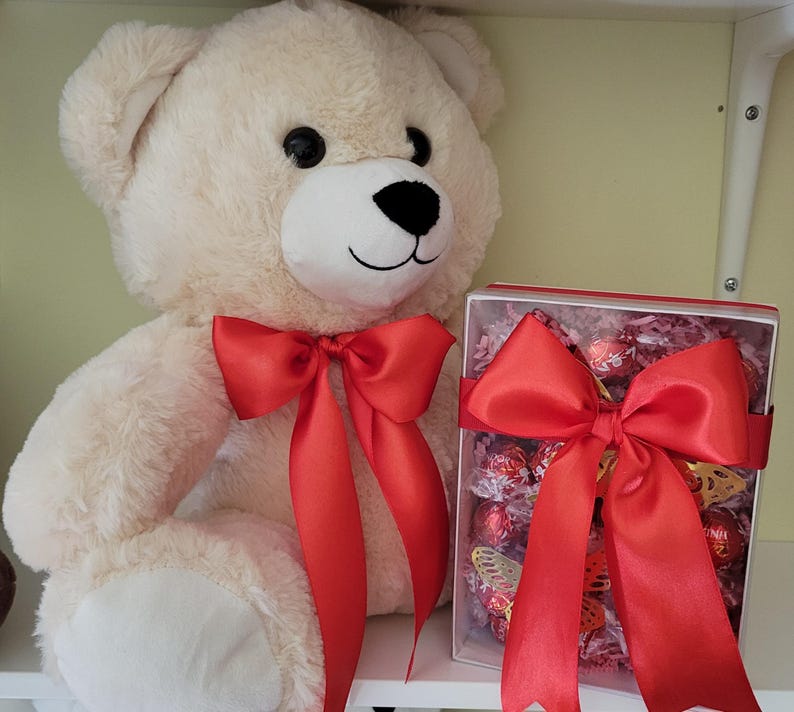 Puede incluir: Un oso de peluche color crema con un lazo de sat&eacute;n rojo alrededor del cuello. Junto al oso hay una caja transparente llena de bombones y un gran lazo de sat&eacute;n rojo. La caja contiene decoraciones de mariposas doradas.