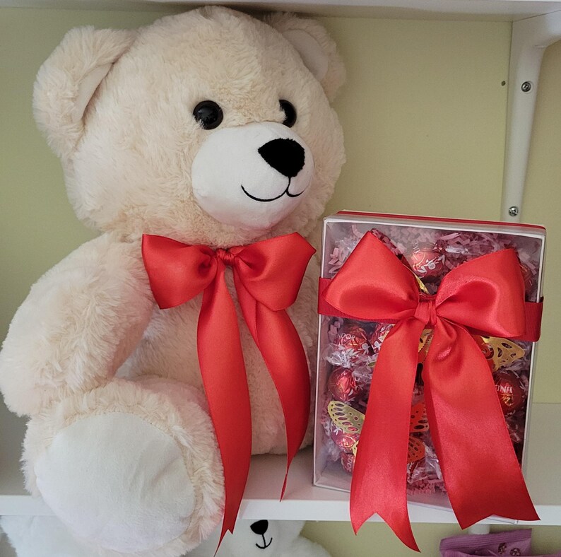 Puede incluir: Un gran oso de peluche color crema con un lazo de sat&eacute;n rojo alrededor del cuello est&aacute; sentado junto a una caja de regalo. La caja est&aacute; llena de bombones y adornada con un lazo rojo a juego. El oso tiene ojos negros y nariz negra.