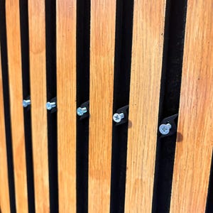 Könnte beinhalten: Nahaufnahme einer Holzlattenwand mit vertikalen, hellbraunen Holzlatten und schwarzen Zwischenräumen. Kleine silberne Schrauben sind sichtbar, die mit schwarzen Halterungen an den Holzlatten befestigt sind. Die Holzmaserung ist sichtbar.
