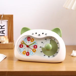 Peut inclure: Minuteur de cuisine blanc en forme de chat avec des oreilles vertes. Le minuteur comporte deux cadrans : un pour l'heure et l'autre pour le compte à rebours. Le cadran de l'horloge a des chiffres colorés. Le minuteur est sur une surface en bois.