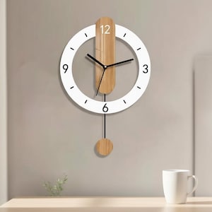 Puede incluir: Un reloj de pared moderno con un marco circular blanco y manecillas negras. El reloj presenta un péndulo de madera y un acento de madera en el centro. Los números se indican con pequeñas rayas negras. Una taza blanca está sobre una superficie de madera.