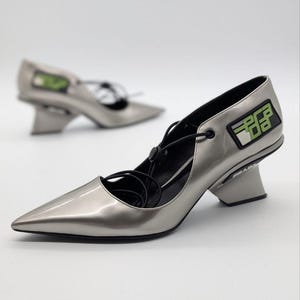 Puede incluir: Zapatos de tacón plateados con punta y diseño de tacón único. Los zapatos presentan cordones negros y un parche con el logo con detalles verdes. Los zapatos de tacón tienen un acabado brillante y una estética moderna.