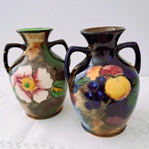 May include: Two decorative ceramic vases. One vase features a floral design with a pink and white flower. The other vase has a fruit design with grapes and a yellow fruit. Both vases have dark handles and rims.