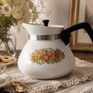 May include: A white enamel teapot featuring a central botanical illustration of oranges, cocoa pods, and yellow flowers. The teapot has a black handle, a black lid knob, and a silver-toned metal band around the upper neck area.