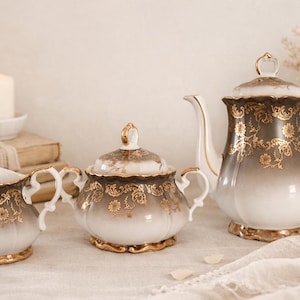 Peut inclure: Service à thé vintage comprenant une théière, un sucrier et un pot à crème. Les pièces en porcelaine blanche présentent un motif floral marron et doré. Une bougie allumée et des fleurs séchées sont en arrière-plan.