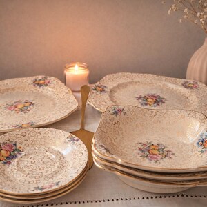 Peut inclure: Ensemble de vaisselle dorée vintage avec un motif floral de fruits. L'ensemble comprend des assiettes, des soucoupes et des bols. Une bougie allumée et un vase décoratif avec des fleurs séchées sont en arrière-plan. Une ustensile de service doré est également présent.