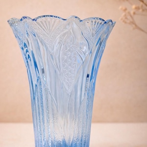 May include: A light blue glass vase with a textured surface and a scalloped rim. The vase features an etched design of leaves and vertical lines, creating a decorative and elegant appearance. The vase is suitable for displaying flowers.