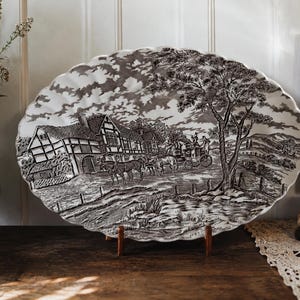 May include: An oval ceramic platter with a scalloped edge featuring a black and white transferware scene of a horse-drawn carriage travelling past a timber-framed house and a large tree in a rural landscape with a cloudy sky and a small pond.