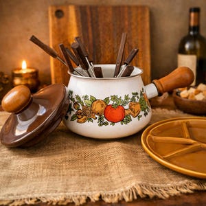 May include: A white fondue pot with a wooden handle and a lid. The pot is decorated with a colorful fruit and vegetable design. Includes fondue forks and a divided serving plate. A wooden cutting board and a bottle of wine are in the background.