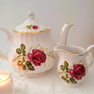 Peut inclure: Service à thé en porcelaine blanche comprenant une théière et un pot à crème, tous deux ornés de motifs floraux de roses rouges et de bordures dorées. La théière a un couvercle et une anse incurvée. Un bougeoir allumé est au premier plan.