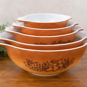 Peut inclure: Un ensemble de quatre bols mélangeurs emboîtables avec un intérieur blanc et un extérieur orange brûlé. Chaque bol présente une illustration au trait brun foncé de raisins et de pommes sur le devant. Les bols ont des poignées intégrées.