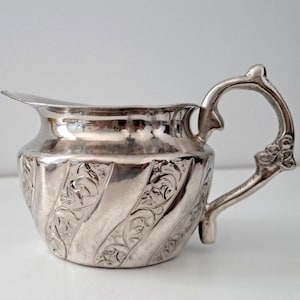Peut inclure: Une crémière argentée avec un bec verseur incurvé et une anse ornée. Le corps présente un motif en relief tourbillonnant de motifs floraux. La crémière est en métal brillant et réfléchissant, probablement de l'argent ou un alliage argenté.
