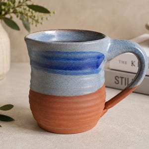 Peut inclure: Une tasse en céramique présentant un design bicolore avec une base texturée en terre cuite non émaillée et une partie supérieure lisse émaillée bleu clair. Une bande horizontale de glaçure bleu cobalt profond entoure le haut.