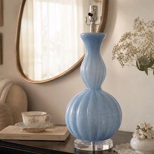 Peut inclure: Une lampe de table en verre bleu clair avec un corps côtelé en forme de gourde et une base circulaire transparente. La lampe présente une douille métallique au sommet, conçue pour recevoir une ampoule, et possède une finition lisse et brillante.