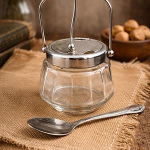 Peut inclure: Un sucrier en verre transparent avec un couvercle et une poignée argentés, et une cuillère assortie, est posé sur une toile de jute. Le bol a un design à facettes. La cuillère a un manche décoratif. Le fond comprend un bol en bois de noix.