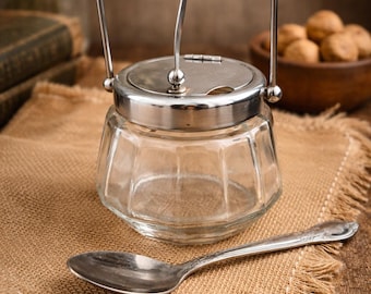 May include: A clear glass sugar bowl with a silver lid and handle, and a matching spoon, sits on a burlap cloth. The bowl has a faceted design. The spoon has a decorative handle. The background includes a wooden bowl of nuts.