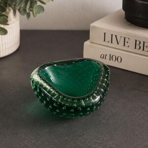 May include: A deep emerald green glass bowl featuring a thick, rounded rim and a distinct bullicante technique with numerous small air bubbles trapped within the glass walls. The bowl has an organic, asymmetrical shape with a smooth, polished finish.
