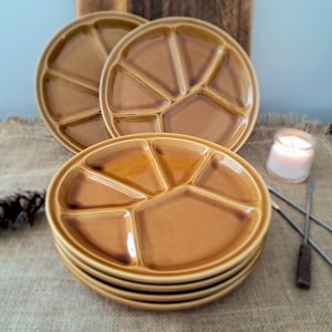 Peut inclure: Une pile de six assiettes divisées en céramique marron avec une finition brillante. Les assiettes sont rondes avec un design segmenté, idéal pour servir des apéritifs ou des collations. Une bougie allumée et des fourchettes à fondue sont à l'arrière-plan.