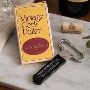 Peut inclure: Un tire-bouchon en métal avec un manche noir portant l'inscription The Christian Brothers 1882 Centennial 1982, accompagné d'une boîte jaune marquée Vintage Cork Puller et The Christian Brothers, ainsi qu'un bouchon avec un blason et le nom de la marque.