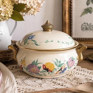 Peut inclure: Une soupière en émail de couleur crème avec un couvercle assorti, ornée d'un motif floral coloré composé de fleurs violettes, jaunes et roses avec des feuilles vertes. La soupière possède deux anses dorées et un bouton doré sur le couvercle.