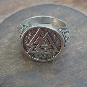 Pode incluir: Um anel de metal prateado com uma face circular apresentando um símbolo Valknut em relevo sobre um fundo escuro e texturizado. A banda do anel exibe padrões de nós intrincados e em relevo ao longo das laterais, conferindo um acabamento detalhado.