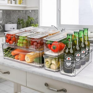 Op de afbeelding: Transparante plastic voedselopslagcontainers gevuld met verse producten, waaronder tomaten, druiven en wortelen. Er zijn ook verschillende groene Heineken-bierflessen te zien, wat wijst op een thema van keuken- of voorraadkastorganisatie.