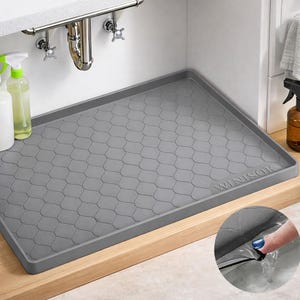 May include: A gray, rectangular sink mat with a textured, fish-scale pattern. The mat is placed under a sink with chrome fixtures and cleaning supplies. The brand name "WINISOK" is visible on the mat.