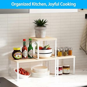 May include: A two-tiered kitchen shelf with white metal legs and light wood shelves. The top shelf holds a potted plant. The shelves display jars, bottles of hot sauce, spices, a bowl of vegetables, and a bowl of tomatoes. The text at the top says "Organized Kitchen, Joyful Cooking."