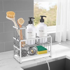 May include: A stainless steel sink organizer holds two white soap dispensers labeled "Hand Soap" and "Dish Soap." It also contains a dish brush, a smaller brush, a stainless steel strainer, a green and yellow sponge, and a gray scrubby.