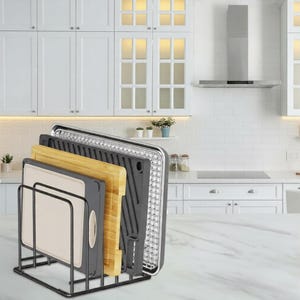 May include: A black metal kitchen organizer holding a variety of cutting boards, a grill pan, and a baking sheet. The cutting boards are made of wood and plastic. The baking sheet has a wire rack. The kitchen is white with white cabinets.