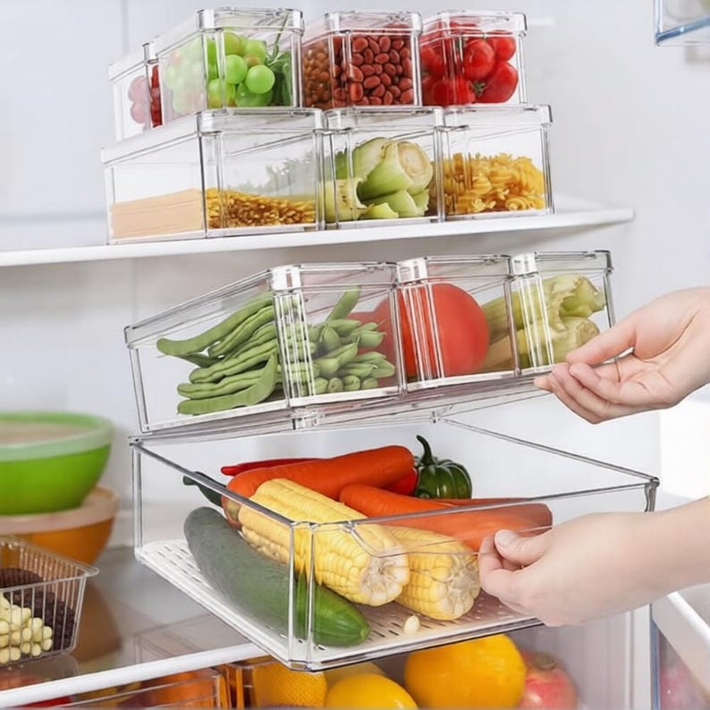 Op de afbeelding: Transparante plastic voedselopslagcontainers gevuld met diverse verse producten, waaronder druiven, bonen, tomaten en ma&iuml;s. De containers zijn gestapeld in een koelkast en bieden een overzicht van georganiseerde voedselopslag.