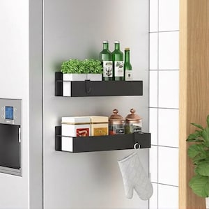 May include: Two black metal shelves mounted on a white surface. The top shelf holds a small potted plant and green bottles. The bottom shelf displays jars, boxes, and a white oven mitt hanging from a hook. Kitchen storage.