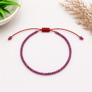 May include: A delicate bracelet featuring a continuous strand of small, faceted, deep magenta-coloured beads. The bracelet is secured with a bright red cord that includes an adjustable sliding knot mechanism and two small beads at the ends.