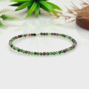 May include: A delicate beaded bracelet featuring small, faceted round stones in alternating shades of forest green and deep magenta. The circular strand is strung on a thin cord and includes a single small, polished silver-toned metal bead.