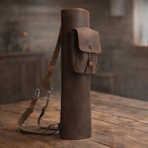 May include: A brown leather quiver with a shoulder strap and a small pouch. The quiver is cylindrical and has a flap closure on the pouch. The leather has a distressed look, and the strap is adjustable. The quiver is designed to hold arrows.