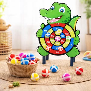 May include: A children's dartboard game with a cartoon alligator design. The target has point values and is held by the alligator. A woven basket contains colorful balls, with some scattered on the floor. The dartboard has a green, yellow, red, and blue color scheme.