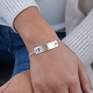 May include: A silver bracelet with a hammered rectangular plate and a small chain. The plate has a stamped design. The bracelet is worn on a wrist, with a neutral-colored sweater and blue jeans in the background.