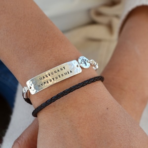 May include: A silver bracelet with the words "MALIGNANT HYPERTHERMIA" engraved on a rectangular plate. A second bracelet is made of dark brown braided leather. The bracelets are worn on a wrist.