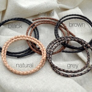 May include: Braided leather cords in various colors: black, camel, brown, natural, and grey. Each cord is coiled, showcasing the texture and color variations. The word "black" is written on the black cord.