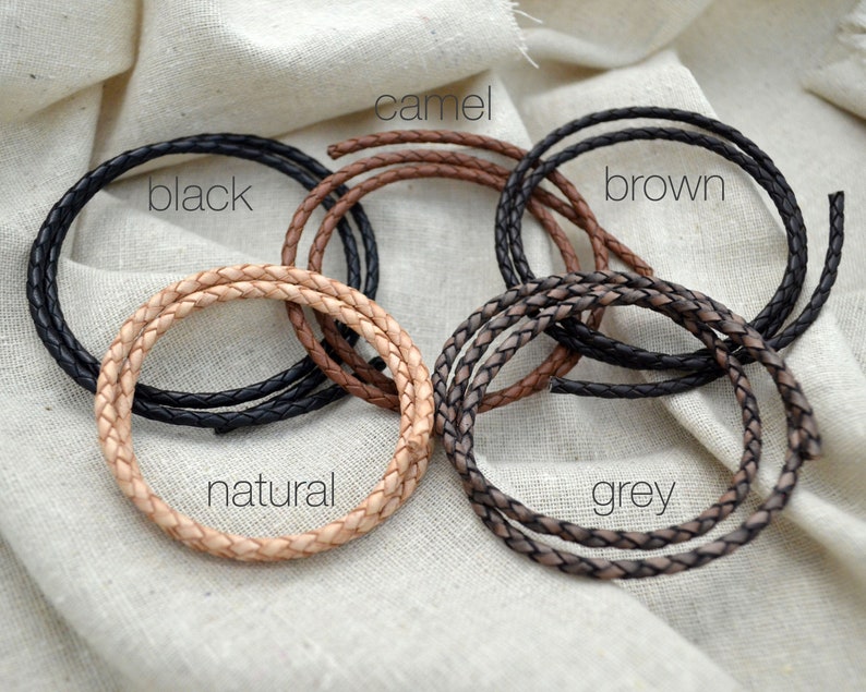 May include: Braided leather cords in various colors: black, camel, brown, natural, and grey. Each cord is coiled, showcasing the texture and color variations. The word "black" is written on the black cord.