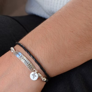 May include: A silver bracelet with the words "TYPE 2 DIABETES" stamped on it, paired with a black leather bracelet. A small charm with a medical symbol hangs from the silver bracelet. The bracelets are worn on a person's wrist.