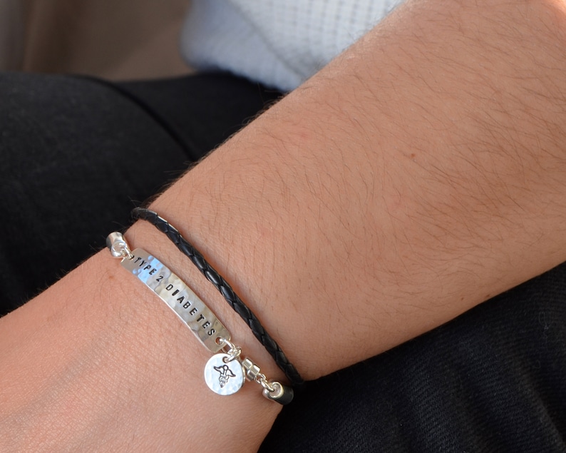 May include: A silver bracelet with the words "TYPE 2 DIABETES" stamped on it, paired with a black leather bracelet. A small charm with a medical symbol hangs from the silver bracelet. The bracelets are worn on a person's wrist.