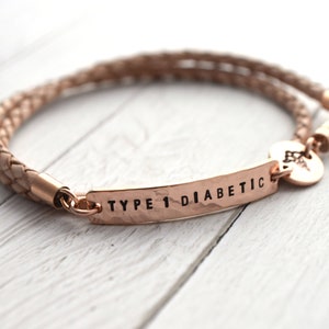 K&ouml;nnte beinhalten: Ein ros&eacute;goldenes medizinische Notfallarmband mit geflochtenem Lederband. Auf der Metallplatte steht "TYPE 1 DIABETIC". Ein kleiner Anh&auml;nger mit einem medizinischen Symbol ist angebracht.