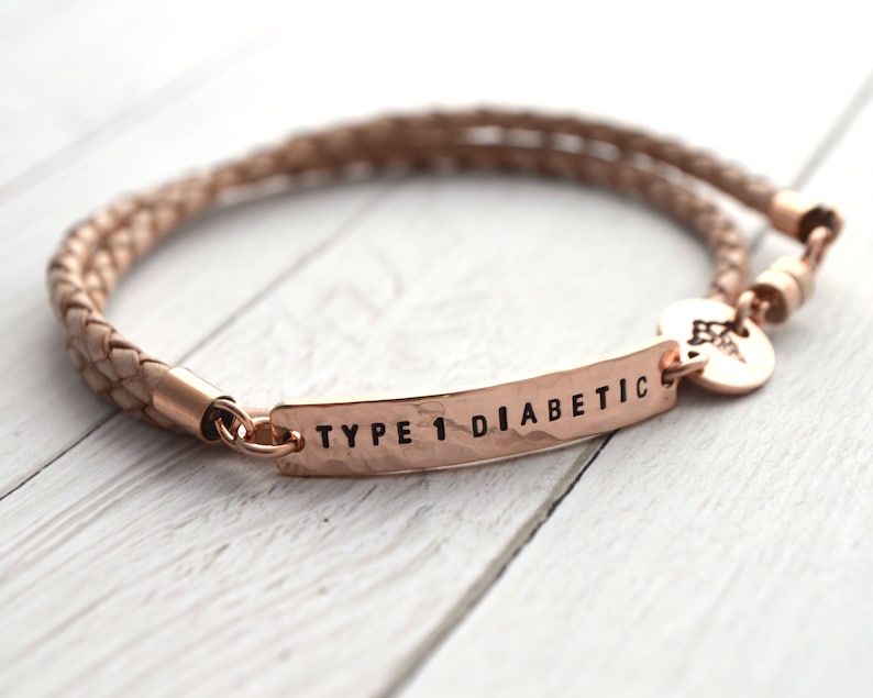 K&ouml;nnte beinhalten: Ein ros&eacute;goldenes medizinische Notfallarmband mit geflochtenem Lederband. Auf der Metallplatte steht "TYPE 1 DIABETIC". Ein kleiner Anh&auml;nger mit einem medizinischen Symbol ist angebracht.