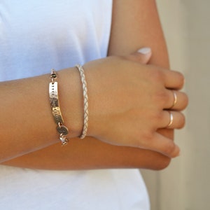 K&ouml;nnte beinhalten: Ein goldfarbenes Armband mit gestempeltem Text und ein geflochtenes Lederarmband werden gezeigt. Das goldene Armband hat eine gebogene rechteckige Platte mit gestempelten Buchstaben und einen kleinen runden Anh&auml;nger. Das geflochtene Armband ist hellbeige.