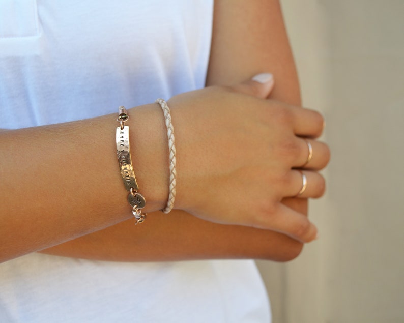 K&ouml;nnte beinhalten: Ein goldfarbenes Armband mit gestempeltem Text und ein geflochtenes Lederarmband werden gezeigt. Das goldene Armband hat eine gebogene rechteckige Platte mit gestempelten Buchstaben und einen kleinen runden Anh&auml;nger. Das geflochtene Armband ist hellbeige.