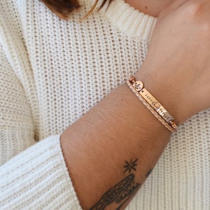 K&ouml;nnte beinhalten: Ein ros&eacute;goldenes Armband mit einem geflochtenen Lederband und einem rechteckigen Anh&auml;nger mit dem eingravierten Wort "DUFF". Das Armband hat auch einen kleinen runden Anh&auml;nger mit einem Kompass-Design. Das Armband wird an einem Handgelenk getragen.